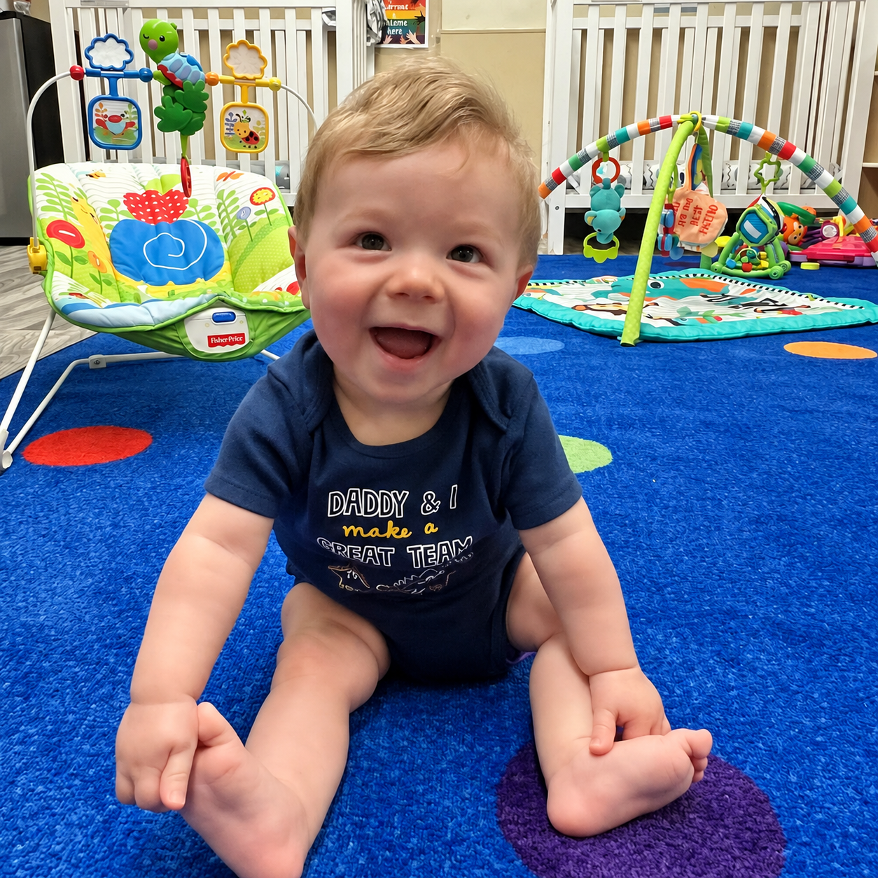A smiling infant sitting on a colorful classroom rug at Kidz-Zona