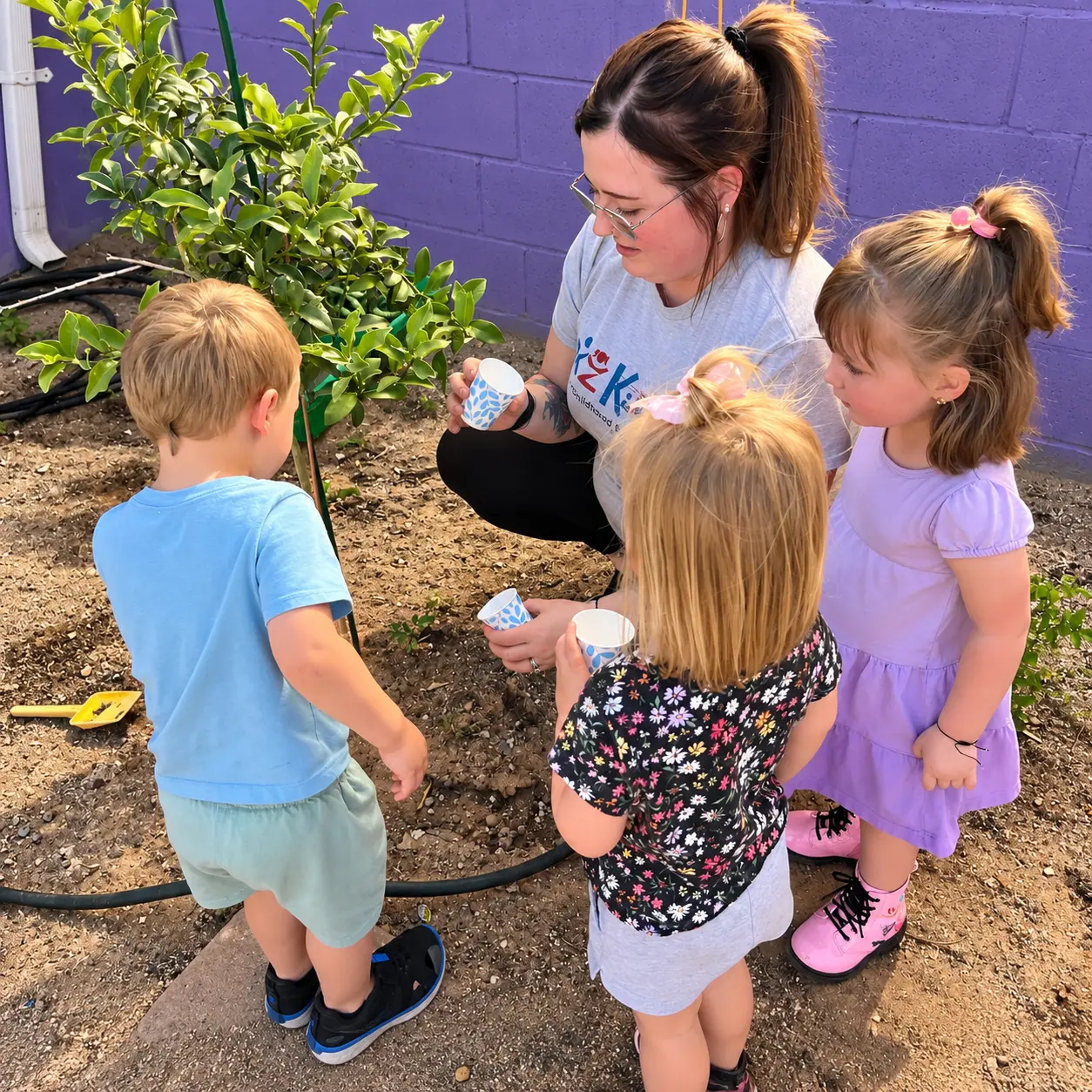 A Kidz-Zona teacher kneeling with three children as they water a newly planted tree in the playground garden