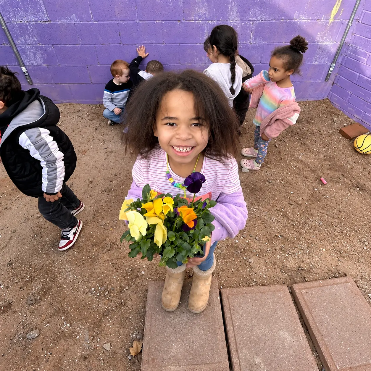 A smiling Pre-K girl holding a pot of colorful flowers on the playground at Kidz-Zona